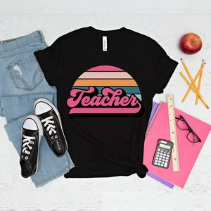 Op de afbeelding: Zwart T-shirt met een retro zonsondergang afbeelding en het woord "Teacher" in roze letters.