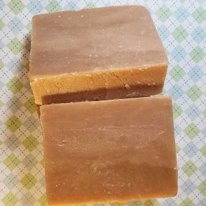 May include: Two bars of handmade soap, one on top of the other. The top bar is a light brown colour with a darker brown stripe in the middle. The bottom bar is a light brown colour. The soap is on a green and white checkered background.