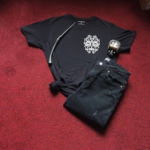 May include: A black t-shirt featuring a white graphic of a skull with intricate details. The shirt is paired with black jeans and a wand. A small figurine of a character with dark hair is also included.