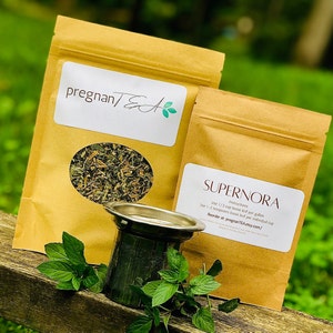 May include: Two brown paper bags with labels that say "pregnanTEA" and "SUPERNORA" with instructions to use 1-2 teaspoons of loose leaf per individual cup or 1/3 cup loose leaf per gallon. The bags are sitting on a wooden surface with a metal tea strainer and green leaves.