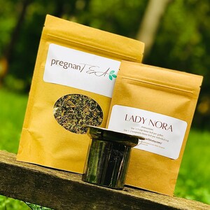 May include: Two brown paper bags with labels that read "pregnanTEA" and "LADY NORA". The LADY NORA bag has instructions for use: "Use 1/3 cup loose leaf per gallon / Use 1-2 teaspoons loose leaf per individual cup". The bags are sitting on a wooden surface next to a silver tea strainer.
