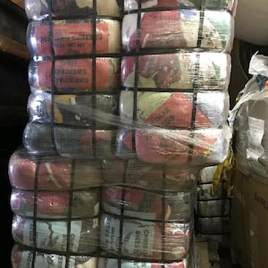 May include: A large, rectangular stack of bundled clothing items wrapped in clear plastic and secured with black straps. The bundles contain various sweaters with visible red, white, green, and grey colors, featuring text labels that read Christmas Sweaters.