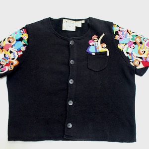 May include: Black button-down shirt with colorful cartoon character sleeves and a small pocket. The sleeves are covered in a pattern of various cartoon figures. A cartoon figure is embroidered on the pocket.