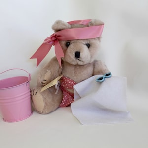 May include: A plush brown teddy bear wearing a pink ribbon headband and a pink and white polka dot swimsuit. The bear is sitting on a white towel with blue sunglasses on top. A pink metal bucket is in the foreground.