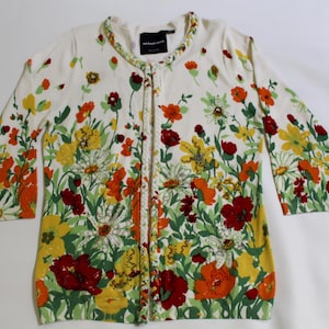 May include: Cream-colored cardigan sweater with a vibrant floral pattern. The design features red, orange, yellow, and white flowers with green leaves. The sweater has a round neckline and long sleeves.