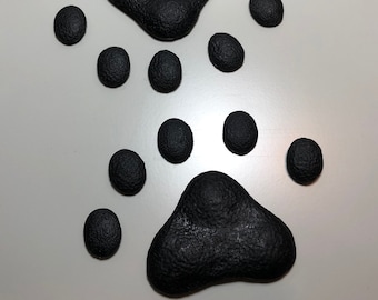 Könnte beinhalten: Schwarze Pfotenabdrücke auf einer weißen Oberfläche. Die Pfotenabdrücke bestehen aus einem strukturierten Material und sind in einem Muster angeordnet, das einer Hundepfote ähnelt.