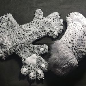 Puede incluir: Un conjunto de patas y cola de animal peludas blancas y grises. Las patas tienen almohadillas blancas y la cola tiene una punta gris.