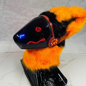 May include: A black and orange furry headpiece with a digital display face. The headpiece has a sleek, futuristic design with orange and black fur accents. The digital display shows red and blue pixelated graphics.