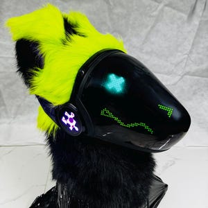 May include: A futuristic, black and neon green furry headpiece. The headpiece features a black visor with green pixelated displays and a neon green and black fur hood. The design includes a purple emblem on the side.