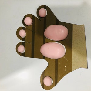May include: A brown cardboard hand with pink foam circles and ovals glued to it. The hand has five fingers and a thumb.