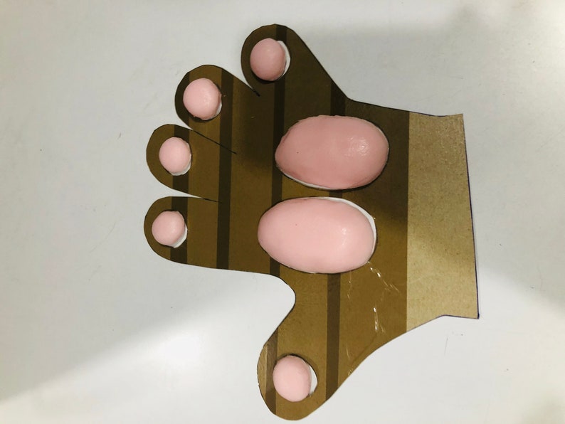 May include: A brown cardboard hand with pink foam circles and ovals glued to it. The hand has five fingers and a thumb.