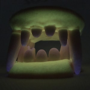 May include: A set of glow-in-the-dark vampire teeth, made of plastic. The teeth are white with a pink hue and have a yellow glow.
