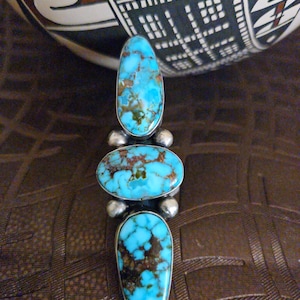 Puede incluir: Un anillo de plata con tres piedras turquesas. Las piedras, de forma ovalada y de lágrima, presentan un color azul vibrante y patrones de matriz marrón. El anillo está sobre un fondo marrón.