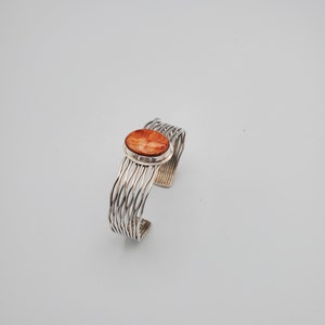May include: A silver cuff bracelet with a woven design. The bracelet features an oval, orange-colored stone set in the center. The metal has a bright, polished finish, creating a striking contrast with the stone.