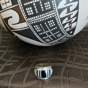 Puede incluir: Un pequeño anillo de plata con un diseño de incrustaciones en blanco y negro. El anillo está frente a un cuenco de cerámica grande, redondo, con dibujos en blanco y negro. El cuenco tiene diseños geométricos y un acento marrón.