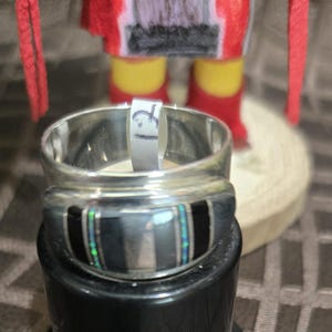 Puede incluir: Un anillo de plata con incrustaciones negras y turquesas. Una etiqueta blanca con el número 12 está dentro del anillo. El anillo está sobre un soporte negro, con una muñeca nativa americana en el fondo.