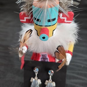 May include: A colorful Native American Kachina doll with a feathered headdress and a white fur collar. The doll is holding feathers and is displayed with a pair of silver earrings with turquoise and red accents.