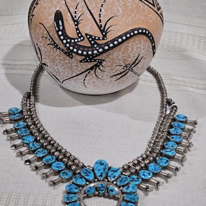 May include: A squash blossom necklace featuring multiple round silver beads and numerous oval-shaped turquoise stones with black matrix. The necklace has a central horseshoe-shaped naja pendant adorned with turquoise stones and silver petal accents.