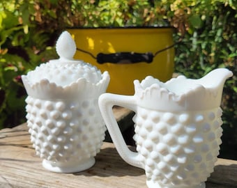 May include: A white milk glass sugar bowl with a lid and a matching creamer pitcher. Both pieces feature a raised hobnail texture, a scalloped rim, and a pedestal base. The sugar bowl has a pointed finial on the lid, and the creamer has a handle.