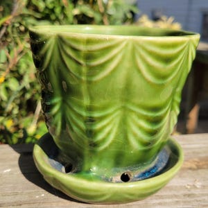 May include: A glossy green ceramic planter featuring a raised, draped fabric-like texture around the exterior. The pot sits securely on a matching circular green saucer that includes visible drainage holes along the base rim.