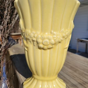 May include: A pale yellow ceramic vase with a fluted rim and a floral garland design around the middle. The vase has a pedestal base and is likely vintage. The vase is outdoors on a wooden surface.