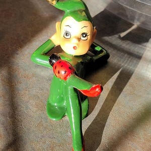 May include: A ceramic figurine of a green elf with a surprised expression. The elf has a gold ball on the tip of its hat and is holding a red and black ladybug. The elf is wearing red shoes.