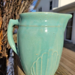 May include: A turquoise ceramic pitcher with a handle and a spout. The pitcher has a textured surface and a decorative fan-like pattern on the lower half. The pitcher is sitting on a wooden surface.