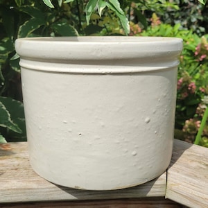 May include: A cylindrical off-white ceramic crock featuring a smooth, glazed finish and a prominent, raised rim around the top edge. The vessel has a simple, uniform design with a slightly textured surface and a flat base, presented in a neutral tone.