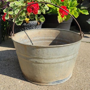May include: A round, galvanized metal bucket with a weathered, light gray finish and a thin, dark metal wire handle attached to the sides. The bucket features a wide, open top and a slightly tapered body with horizontal ridges along the exterior surface.
