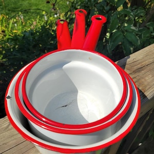 May include: A set of three vintage enamelware pots with bright red handles and rims. The pots are white with a gray exterior and are nested together. The background features green foliage and a wooden surface.