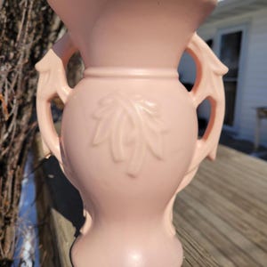 May include: A pale pink ceramic vase with two handles and a flared top. The vase features a leaf design on the front and is sitting on a wooden surface. The vase is likely vintage and suitable for displaying flowers or as a decorative item.