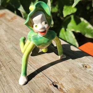 May include: A ceramic figurine of a small creature with a pale face, red lips, and green eyes, wearing a green leaf-shaped hat and collar. The figure has long, slender green limbs with white hands and feet, posed in a crawling position.