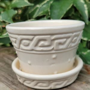 May include: A cream-colored ceramic plant pot with a matching saucer. The pot features a raised Greek key pattern around the upper rim and small, evenly spaced raised dots around the lower body. The finish is smooth and glossy throughout.