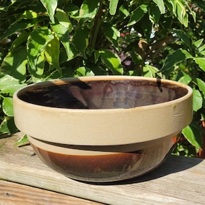 May include: A round ceramic bowl featuring a two-tone design with a smooth, matte beige upper rim and a glossy, dark brown glazed lower section. The interior of the bowl is finished with a deep, dark brown glaze that contrasts with the exterior.