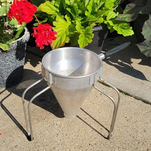 May include: A silver-colored metal food mill or conical strainer featuring a wide circular top, a tapered mesh body, and three sturdy metal legs for stability. It includes a single black handle extending from the side of the rim.
