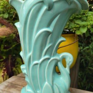 May include: A light turquoise ceramic vase featuring a sculpted swan at the base with stylized, sweeping feather-like patterns extending upward to form the rim. The piece has a glossy finish and a wide, flared opening with a curved silhouette.