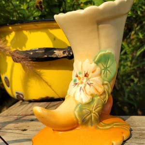 May include: A cream-colored ceramic cornucopia vase featuring a raised relief design of a pale yellow flower with green leaves. The base is a scalloped, vibrant orange oval shape, and the top rim of the vase also has a scalloped edge design.