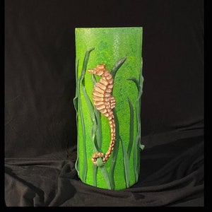 May include: A cylindrical, green decorative vase with a textured surface. A detailed, brown seahorse is affixed to the front, surrounded by green seaweed-like accents. The vase is approximately 10 inches tall.