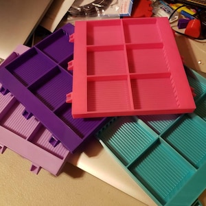 May include: Three plastic storage trays with compartments. The top tray is pink, the middle tray is purple, and the bottom tray is teal.