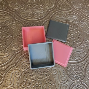 May include: Three square boxes, one grey and two pink, are stacked on top of each other. The boxes are made of a textured material and have a slightly raised edge.