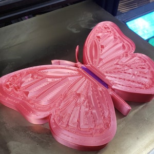 May include: A pink 3D printed butterfly with intricate details. The butterfly is sitting on a grey surface.
