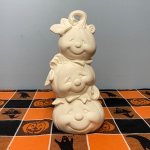 May include: A stack of three white ceramic pumpkins with cute faces. The pumpkins are stacked on top of each other, with the largest pumpkin on the bottom and the smallest pumpkin on top. The pumpkins are all painted white and have a smooth finish.