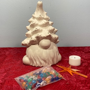 May include: A ceramic gnome-shaped Christmas tree with a round face and beard. The tree has a textured surface and is accompanied by a small candle, a star-shaped ornament, and a bag of colorful lights. The items are on a red surface.