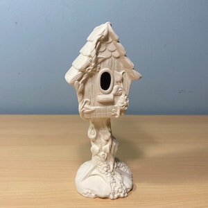 May include: White ceramic birdhouse with a detailed design of vines and leaves. The birdhouse has a round opening for birds to enter and a base with a tree trunk design.
