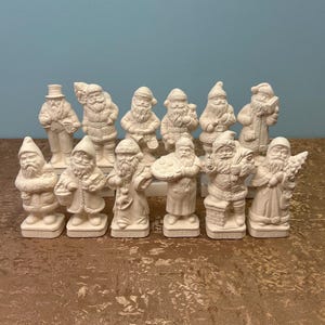 May include: A collection of twelve white ceramic Santa Claus figurines in various poses. Some Santas are holding gifts, while others are carrying sacks or books. The figurines are arranged on a brown surface against a blue background.