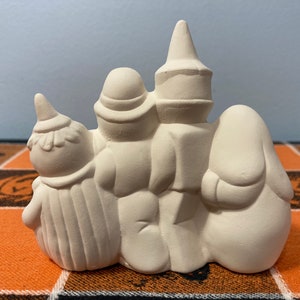 Puede incluir: Una decoraci&oacute;n de Halloween de cer&aacute;mica con tres figuras. Las figuras est&aacute;n dispuestas en fila, con una bruja, un fantasma y una figura con un sombrero alto. Las figuras no est&aacute;n pintadas y listas para personalizar. La decoraci&oacute;n est&aacute; sobre un mantel con tem&aacute;tica de Halloween.