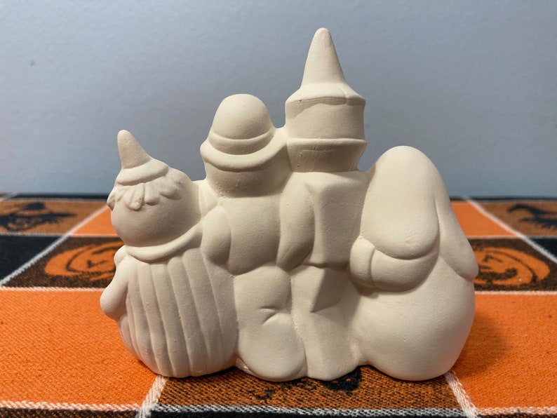 Puede incluir: Una decoraci&oacute;n de Halloween de cer&aacute;mica con tres figuras. Las figuras est&aacute;n dispuestas en fila, con una bruja, un fantasma y una figura con un sombrero alto. Las figuras no est&aacute;n pintadas y listas para personalizar. La decoraci&oacute;n est&aacute; sobre un mantel con tem&aacute;tica de Halloween.