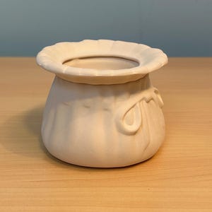 May include: A matte white ceramic vessel shaped like a small fabric pouch with a gathered, ruffled rim and a sculpted bow detail on the side. The container has a wide, circular opening and a rounded base, featuring a smooth, unglazed finish.