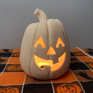 May include: A white ceramic pumpkin with a carved jack-o'-lantern face. The pumpkin is lit from inside, creating a warm glow.