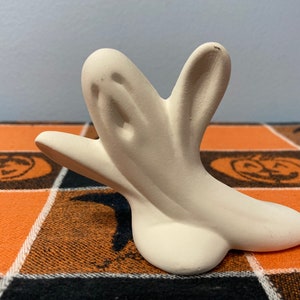 Puede incluir: Una figura de fantasma de cer&aacute;mica de color crema claro. El fantasma tiene una cara simple y amigable y los brazos extendidos. La figura est&aacute; sobre un mantel a cuadros naranja y negro con tem&aacute;tica de Halloween.